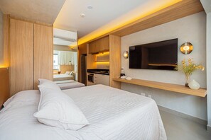 A modern hotel room with a large bed, a flat-screen TV mounted on the wall, a small kitchen area, and a bathroom visible through an open door. - Flat c/ Piscina e Academia na Chácara Klabin (São Paulo)