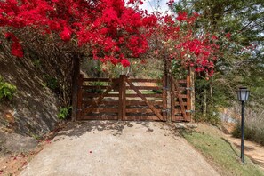 A gated entrance with a concrete path leading through it, surrounded by lush greenery and red flowering plants. - Casa com fogão a lenha para 7 | Vale das Videiras (Itaipava, Petropolis)
