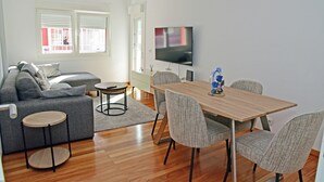 A modern living room with a grey sofa, a round coffee table, a dining table with four chairs, a flat-screen TV mounted on the wall, and a wooden floor.