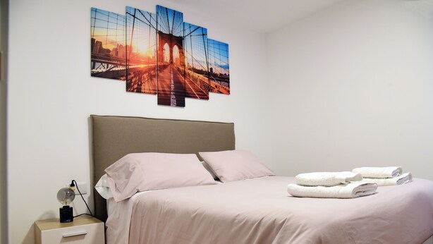 A neatly made bed with a headboard, a bedside table with a lamp, and a wall-mounted artwork of a bridge.