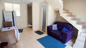 Un salon avec un canapé violet, une table à manger recouverte d’une nappe en dentelle, un escalier et une porte donnant sur une salle de bain.