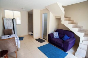 A living room with a purple sofa, a dining table with a lace tablecloth, a staircase, and a door leading to a bathroom. - Casa Pet Friendly com Varanda e Churrasqueira (Rio das Ostras)