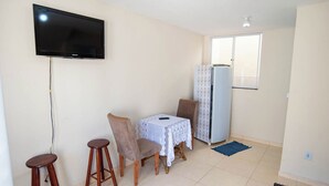 Un espace de vie compact doté d’un téléviseur à écran plat fixé au mur, d’une petite table à manger recouverte d’une nappe en dentelle, de deux chaises, d’un réfrigérateur et d’une fenêtre laissant entrer la lumière naturelle.