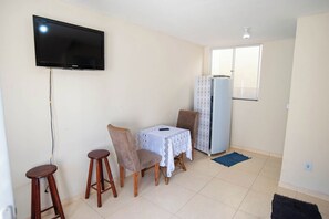 A compact living space with a flat-screen TV mounted on the wall, a small dining table with a lace tablecloth, two chairs, a refrigerator, and a window providing natural light. - Casa Pet Friendly com Varanda e Churrasqueira (Rio das Ostras)