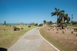 A paved pathway leading to a beach with palm trees and a small statue. - Casa Pet Friendly com Varanda e Churrasqueira (Rio das Ostras)