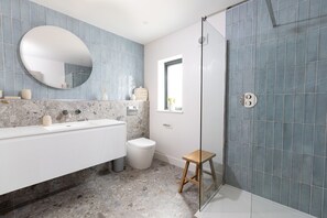 Een badkamer met een witte wastafel, een ronde spiegel, een douche met glazen deur en een klein houten krukje.
