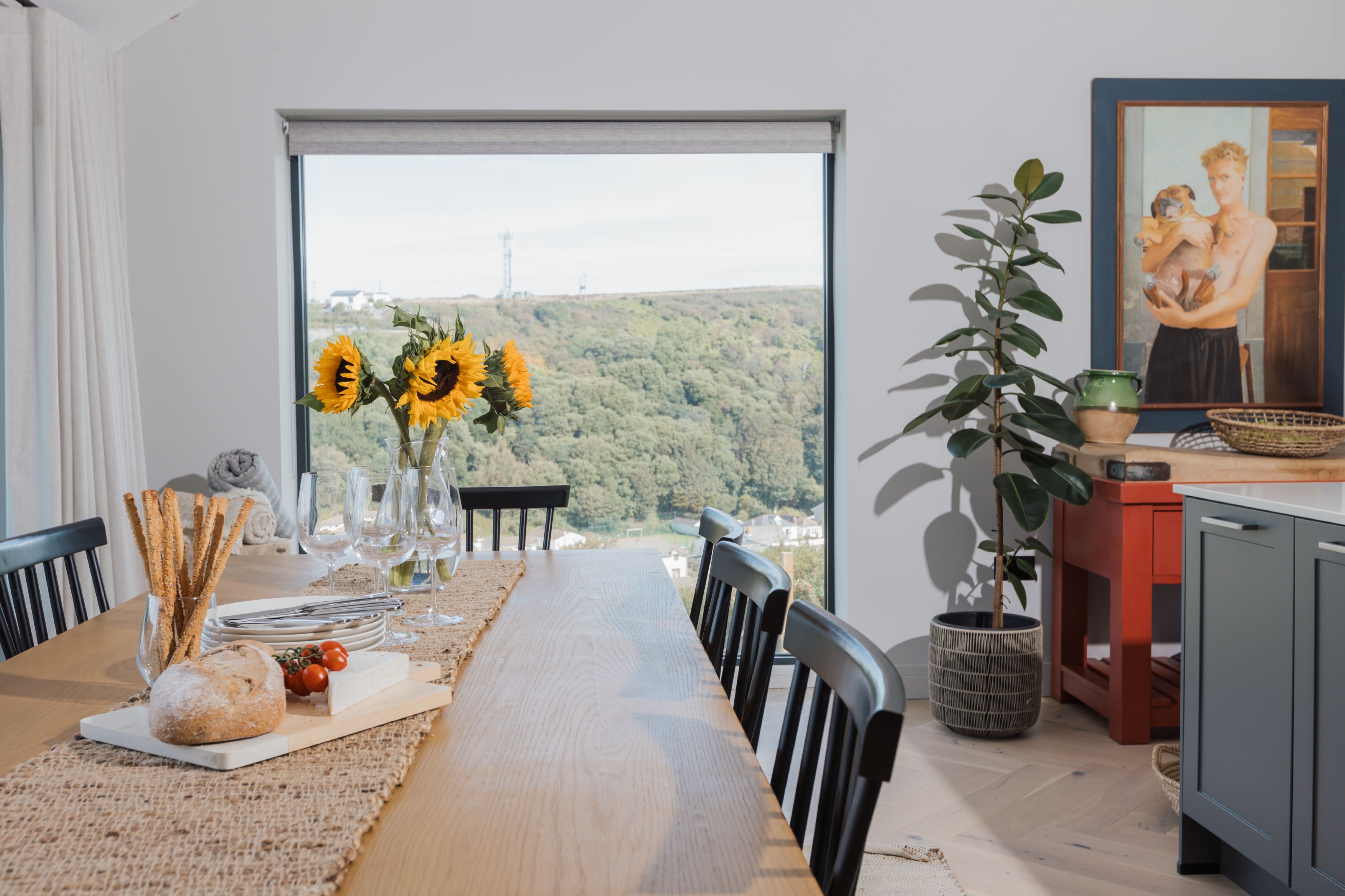 A dining area with a view of a hilly landscape, a table set with plates, glasses, and a vase of sunflowers.
