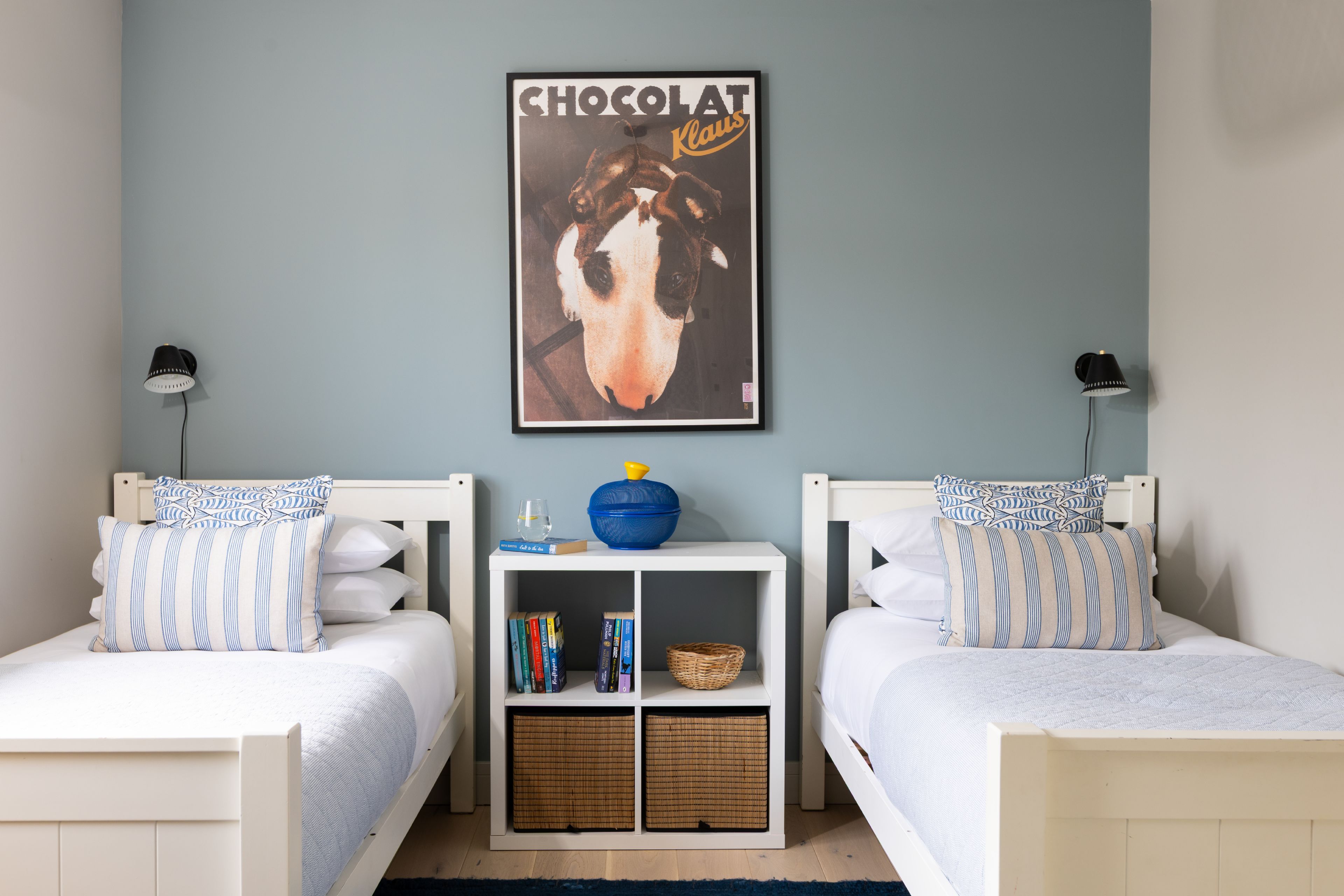 A bedroom with two beds, a nightstand with books and a blue bowl, and a poster on the wall.