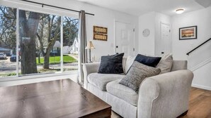 A living room with a sofa, a coffee table, a window with curtains, a door, and a wall with a framed picture and a sign.