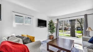 A modern living room with a flat-screen TV, a large window with blinds, a potted plant, and a coffee table.