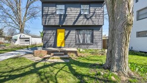 A two-story house with a yellow front door, a brick foundation, and a large tree in front.