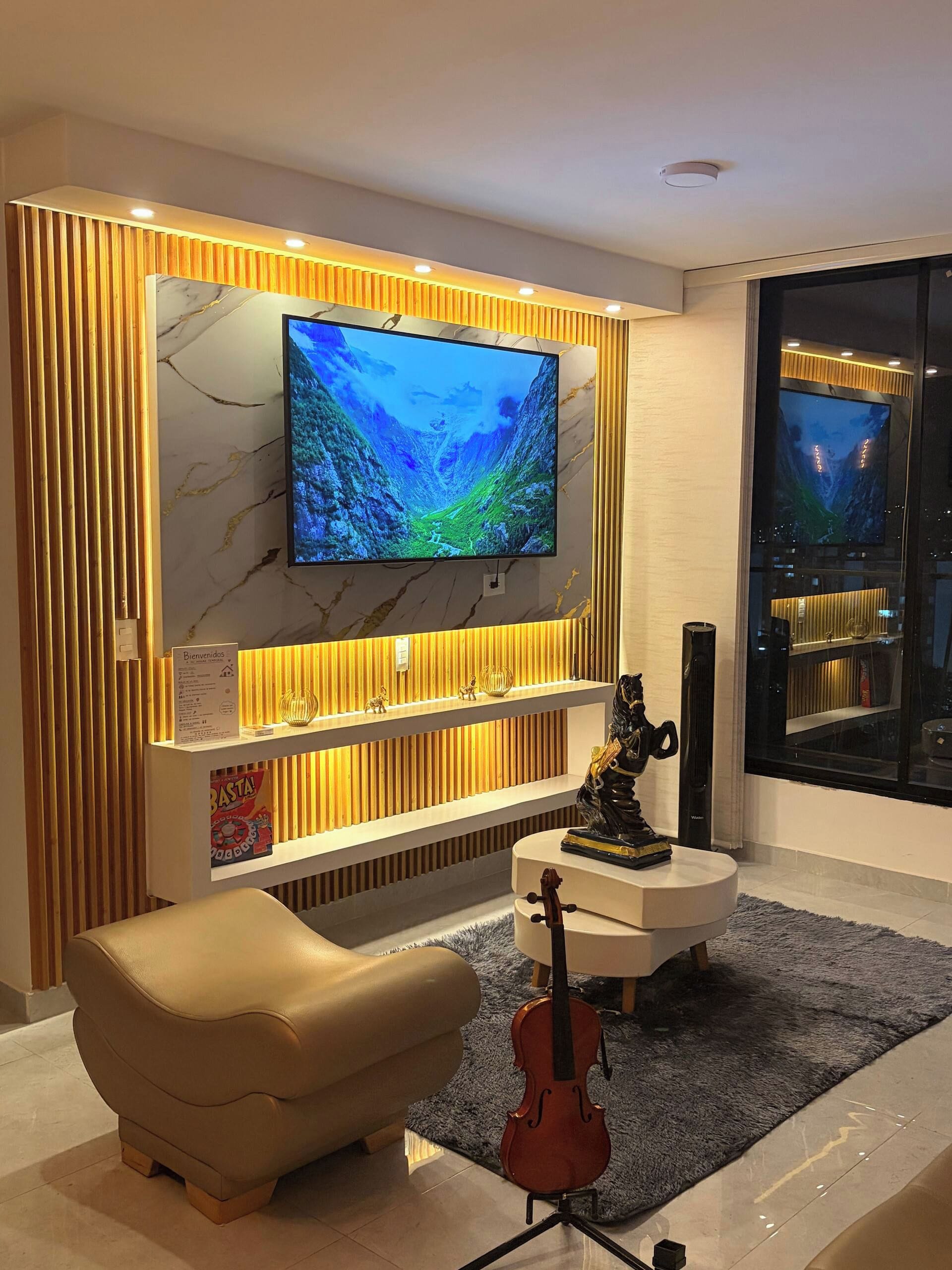 A modern living room with a flat-screen TV mounted on a marble wall, a comfortable sofa, a small round coffee table, and a violin.