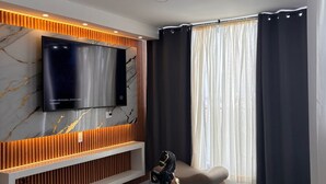 Ett modernt vardagsrum med en platt-TV som hänger på en vägg klädd i marmor, en dekorativ häststaty, en cello och ett fönster med mörka gardiner.