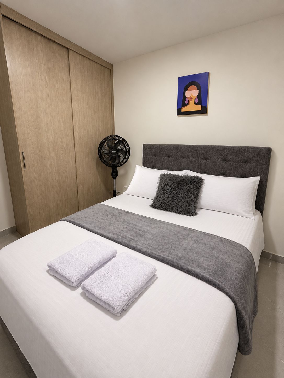 Una cama bien hecha con sábanas blancas y dos toallas dobladas, un cabecero gris, un plaid gris, un ventilador de pie negro y un armario de madera.