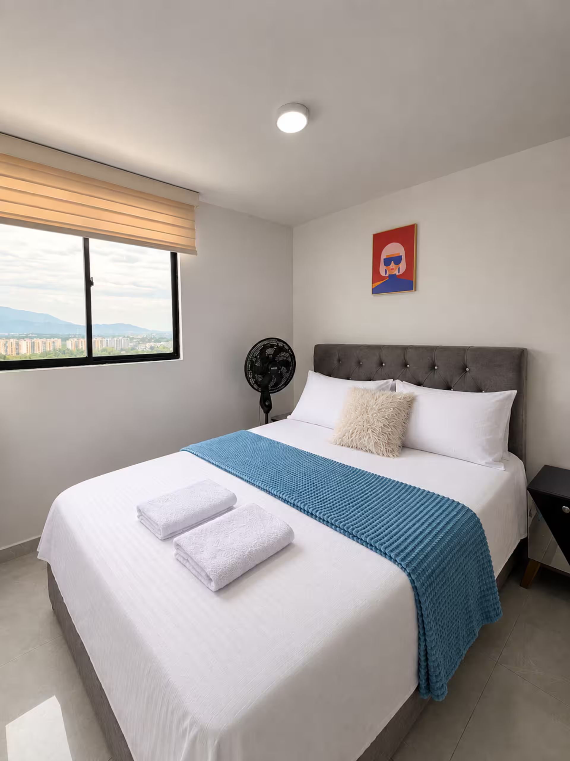 Ein Hotelzimmer mit Bett, Fenster, Ventilator und einem Gemälde an der Wand.