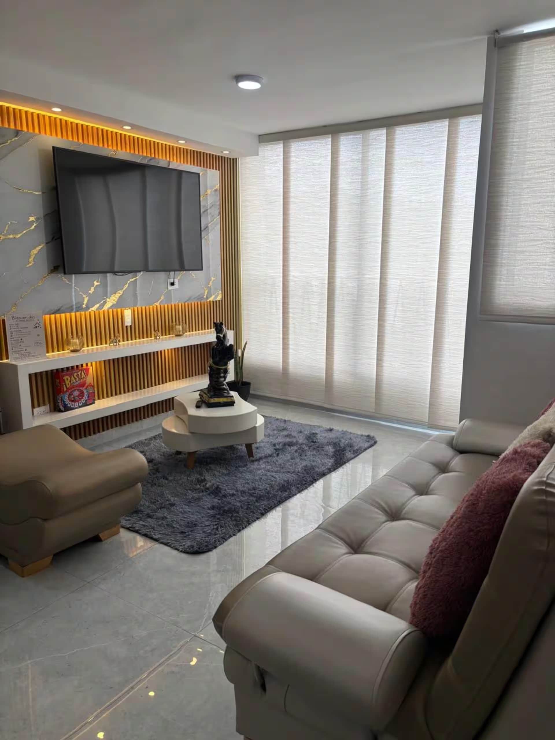 A modern living room with a flat-screen TV mounted on a wall with a marble-like pattern, a sofa, a coffee table, and a large window with curtains.