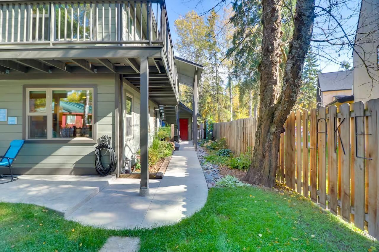 A concrete walkway leads beneath the upper balcony alongside the yard and fence.