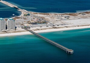 An aerial view of a coastal area with a long pier extending into the water, a sandy beach, and several buildings including a hotel with multiple floors. - Sandy Paws-Pet Friendly Coastal Cottage (Navarre)