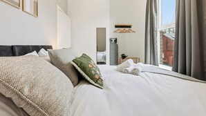 Una cama bien hecha con almohadas, una mesita de noche con hervidor y una ventana con cortinas.