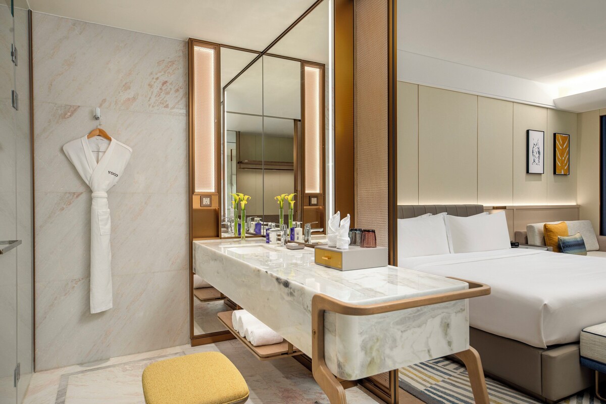 Habitación de hotel moderna con tocador de mármol, un espejo grande, una cama con ropa blanca y un pequeño rincón de descanso.