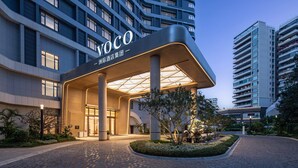 Une entrée d'hôtel moderne, avec une enseigne « Voco » bien visible, un espace d'entrée abrité et un jardin aménagé.