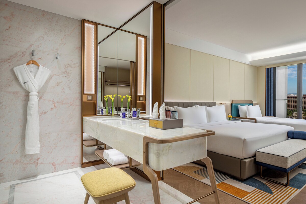 Habitación de hotel con pared de mármol, un espejo grande, tocador con artículos de aseo, dos camas y vistas a la ciudad.