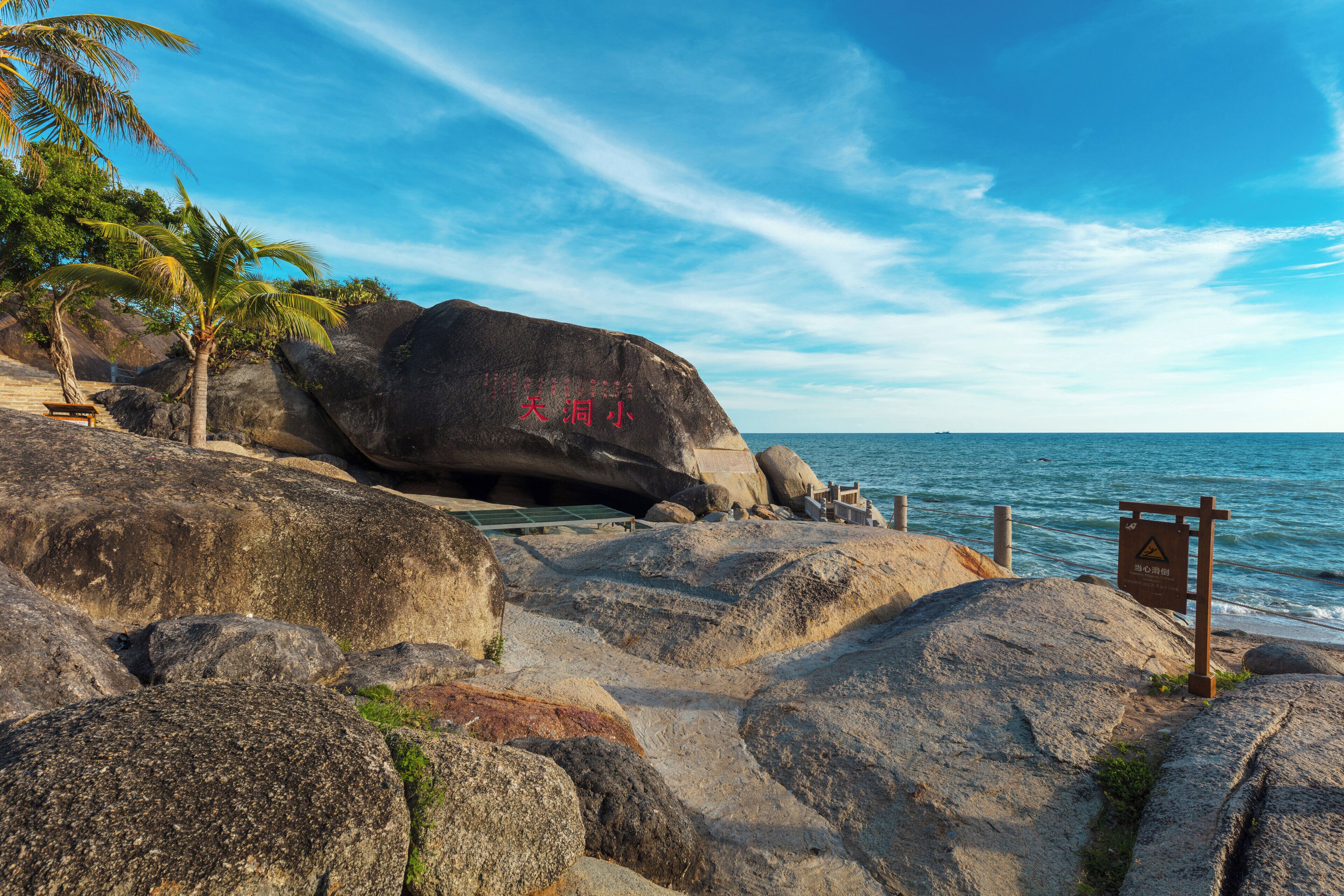 A rocky beach with large boulders, a palm tree, and a bench.