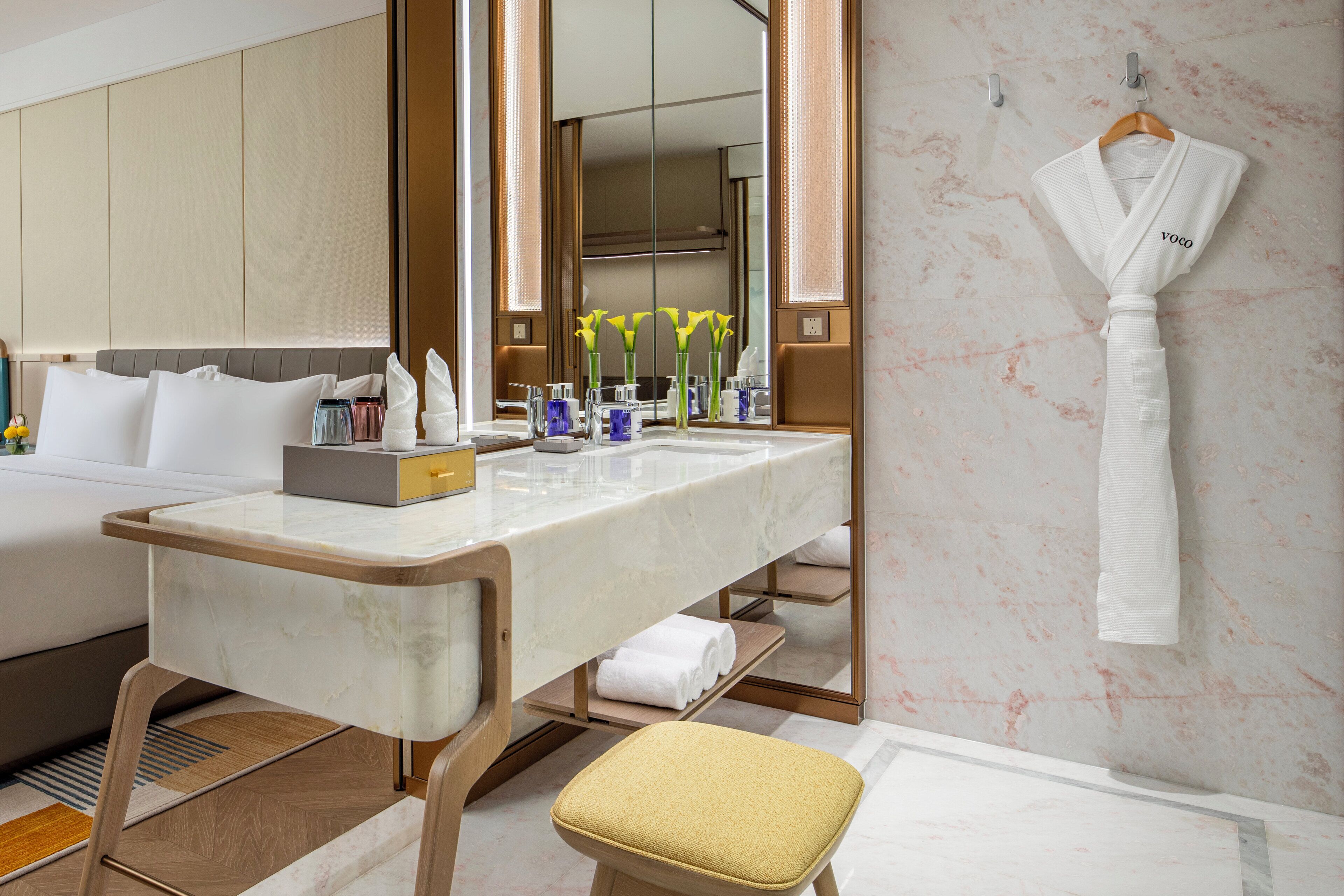 Habitación de hotel con tocador de mármol, cama con ropa blanca, mesita de noche con artículos de aseo y una bata colgada.