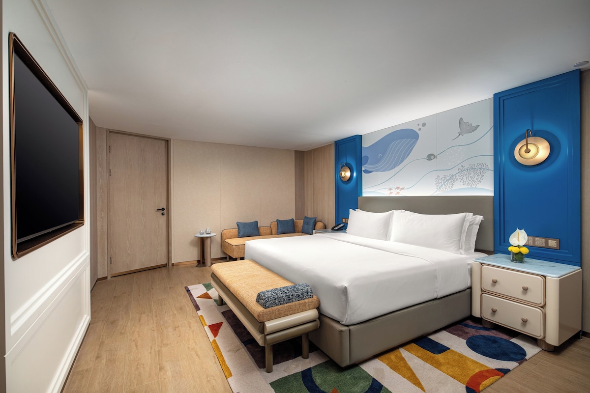 Une chambre d’hôtel dotée d’un grand lit, de tables de chevet, d’un petit coin salon, d’un téléviseur à écran plat et d’un mur orné d’une œuvre d’art sur le thème de la mer.