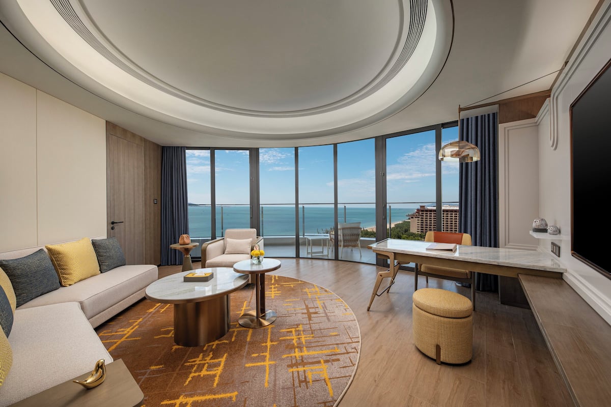 A modern hotel room with a large window offering a view of the ocean, a sofa, a dining table, and a flat-screen TV.