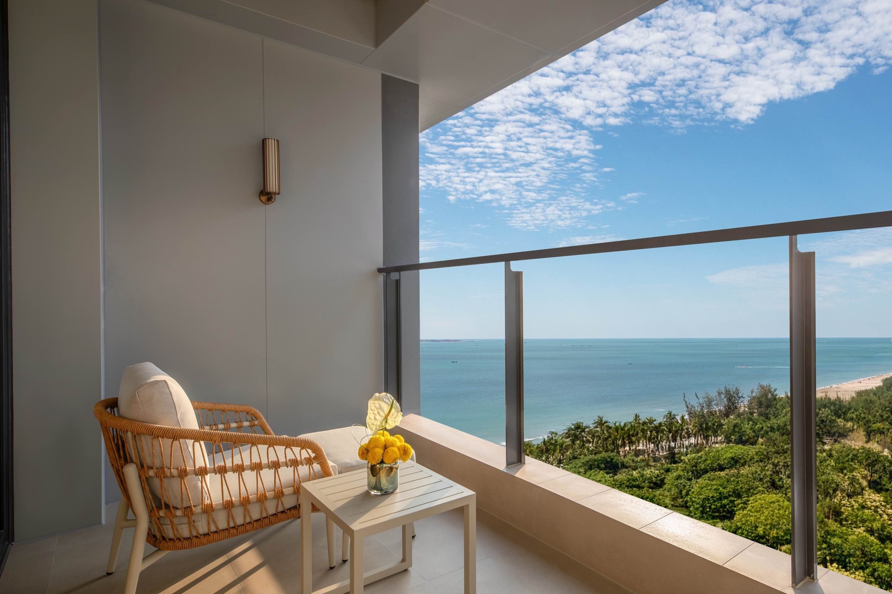 A balcony with a wicker chair, a small table, and a vase of yellow flowers. A large window offers a view of the ocean and a beach.