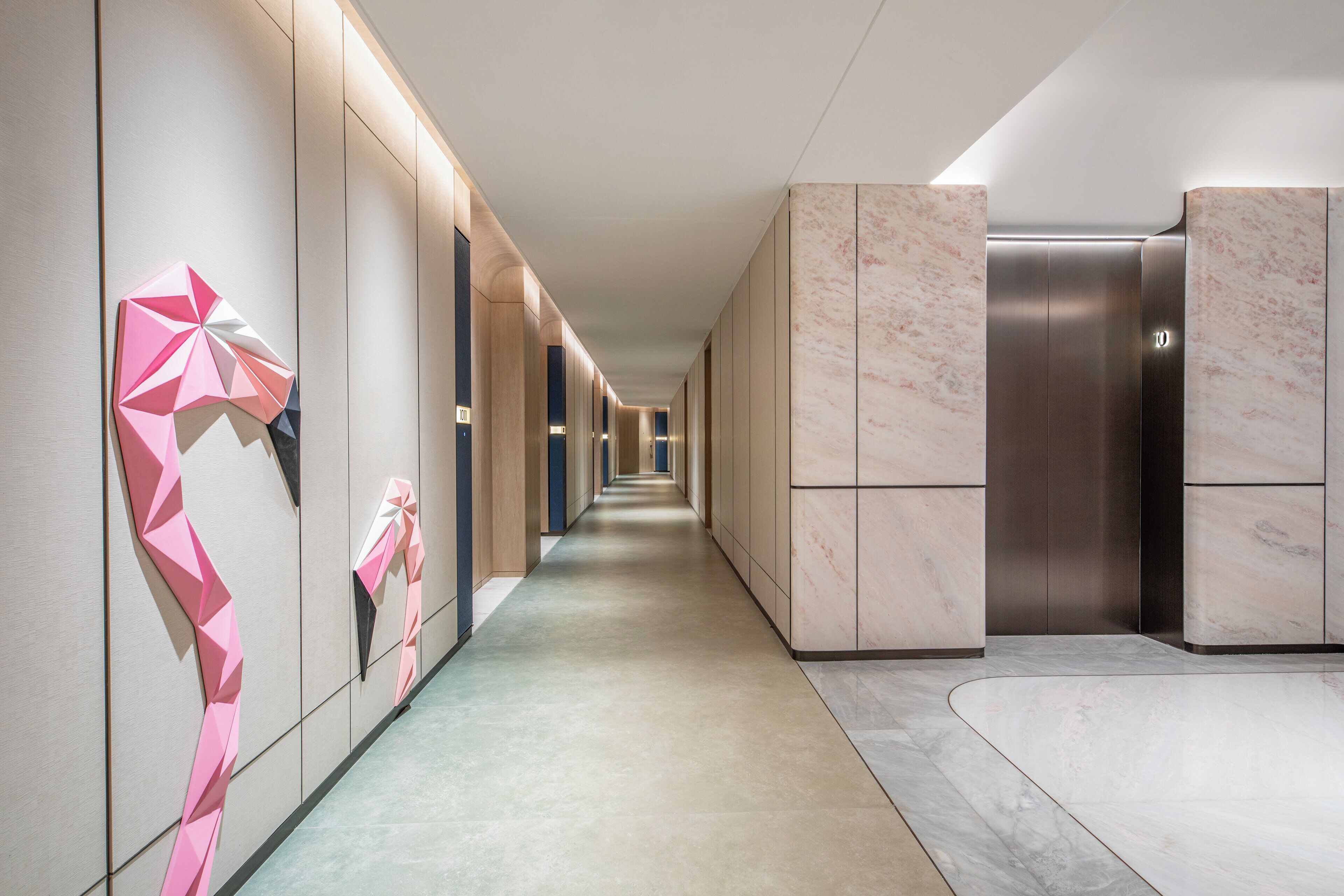 Un corridoio moderno con una lunga galleria, un ascensore metallico e un muro decorato con arte geometrica.