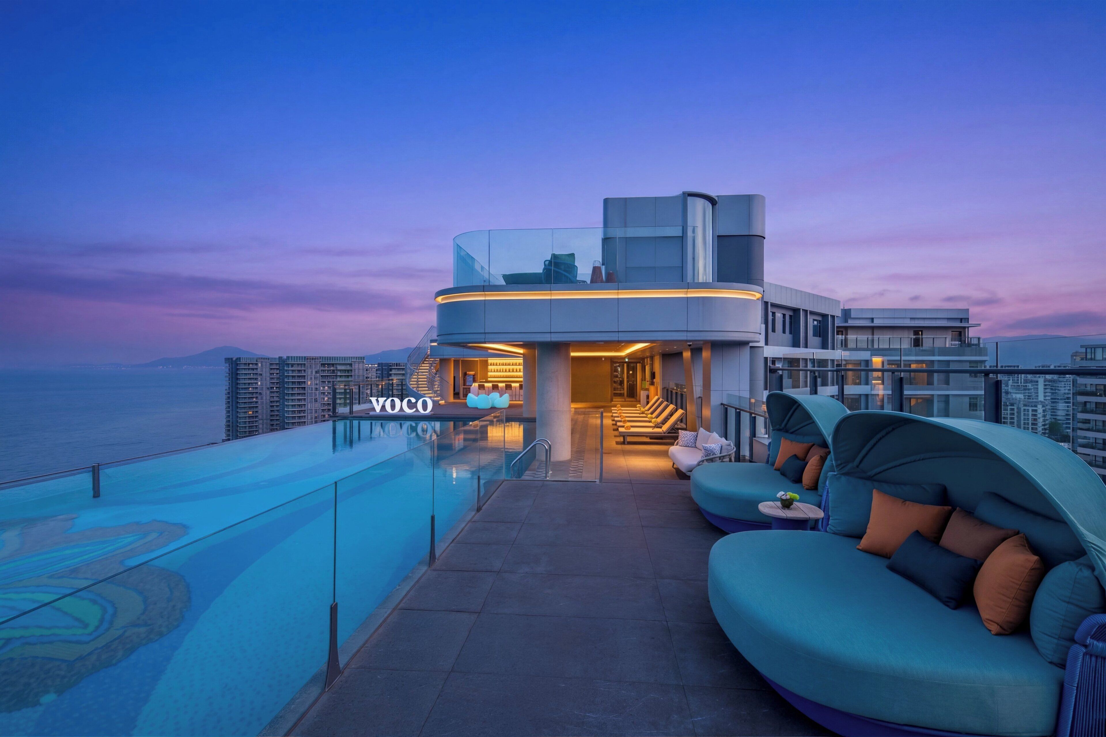 A modern hotel with a glass infinity pool, outdoor seating, and a view of the ocean.