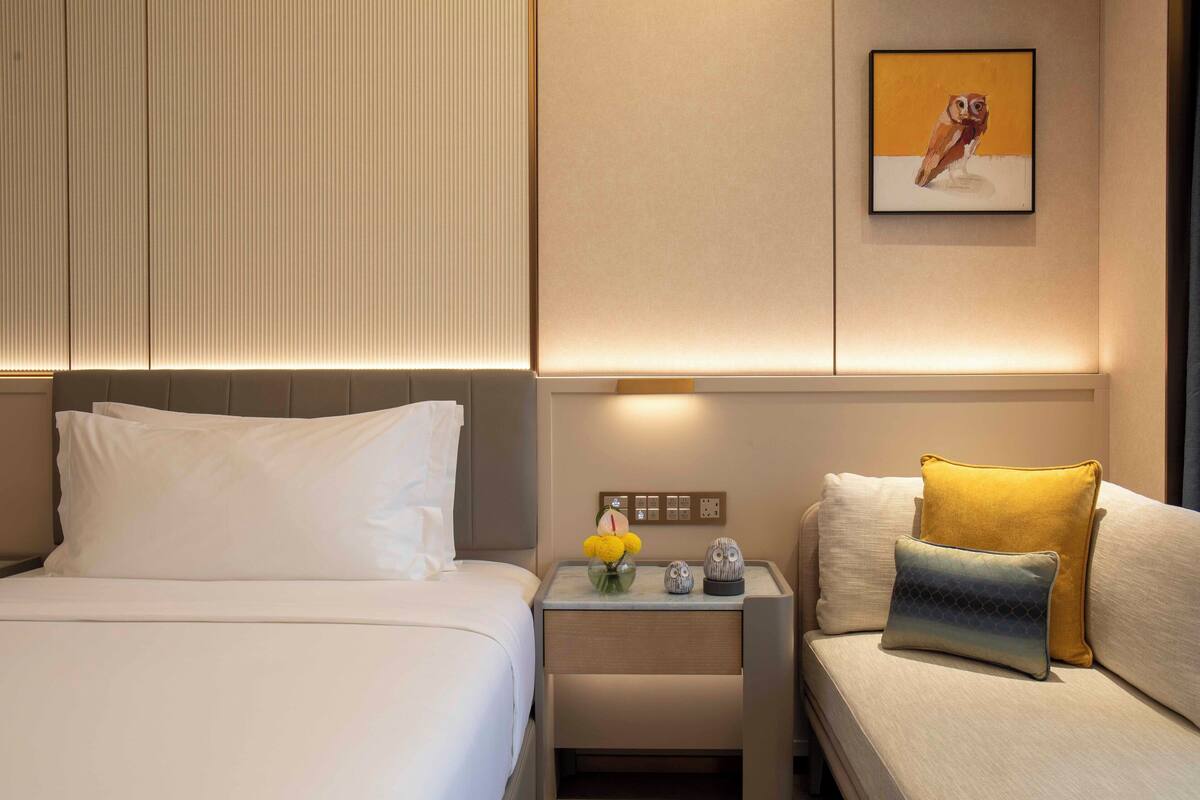 Une chambre d’hôtel avec un lit, une table de chevet ornée d’un vase de fleurs et un canapé avec des coussins.