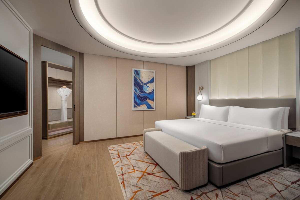 Una habitación de hotel moderna con una cama grande, un televisor, un cuadro y un baño con espejo y toallero.