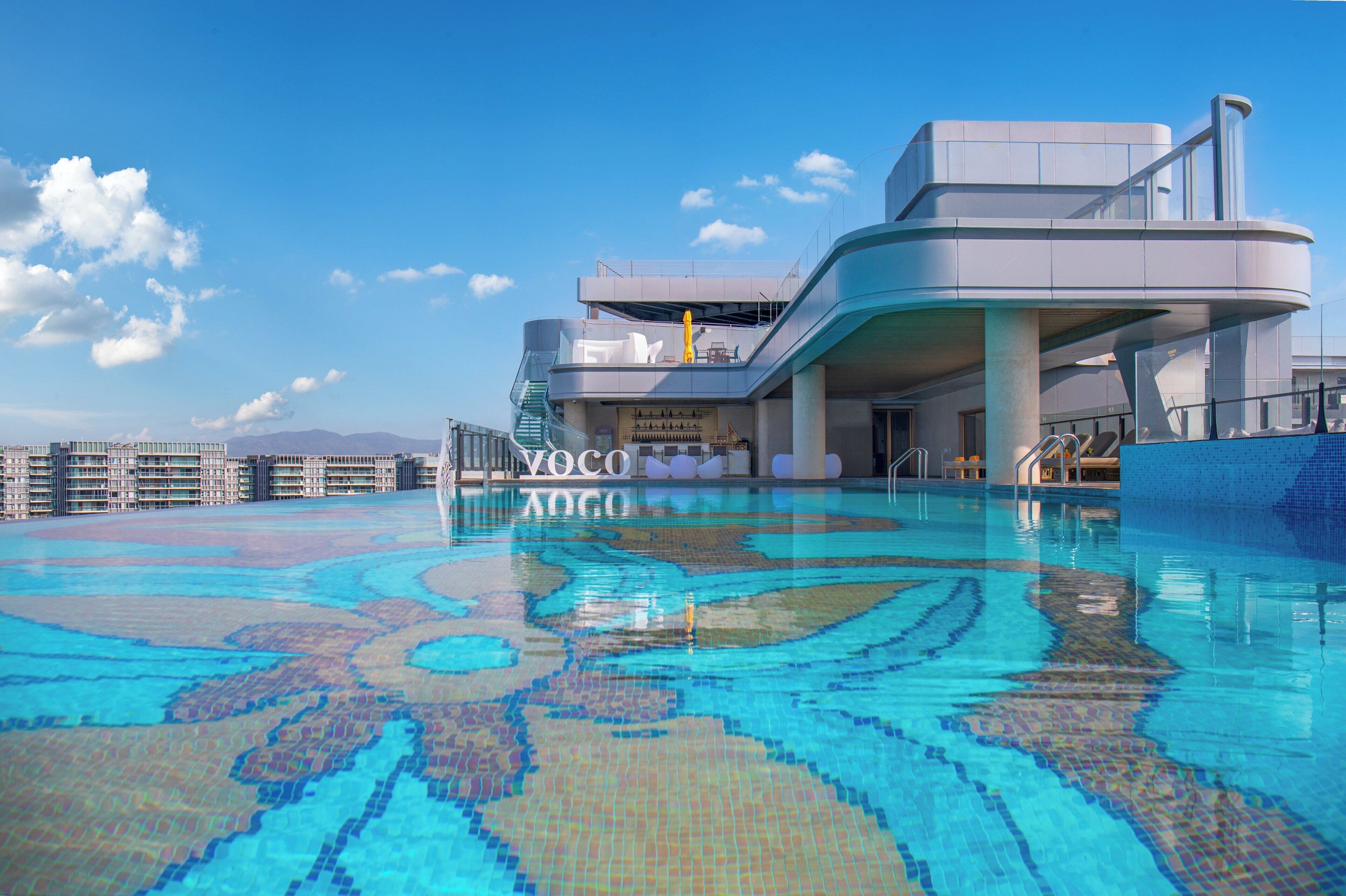 Um hotel moderno com uma grande piscina exterior, área coberta com assentos e vista para montanhas ao longe.