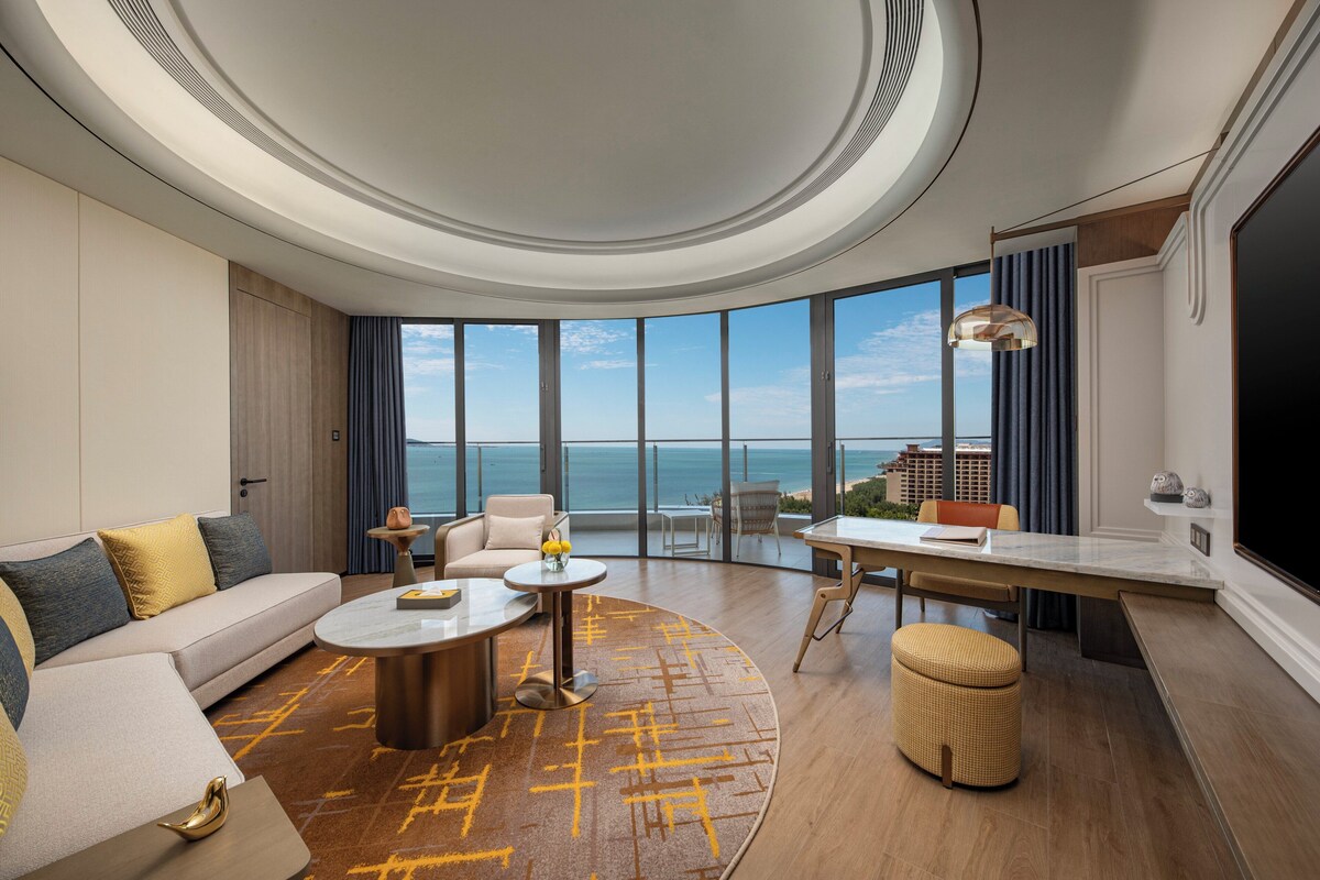 Una habitación de hotel moderna con un gran ventanal que ofrece vistas al océano, un sofá, una mesa de comedor y un televisor de pantalla plana.