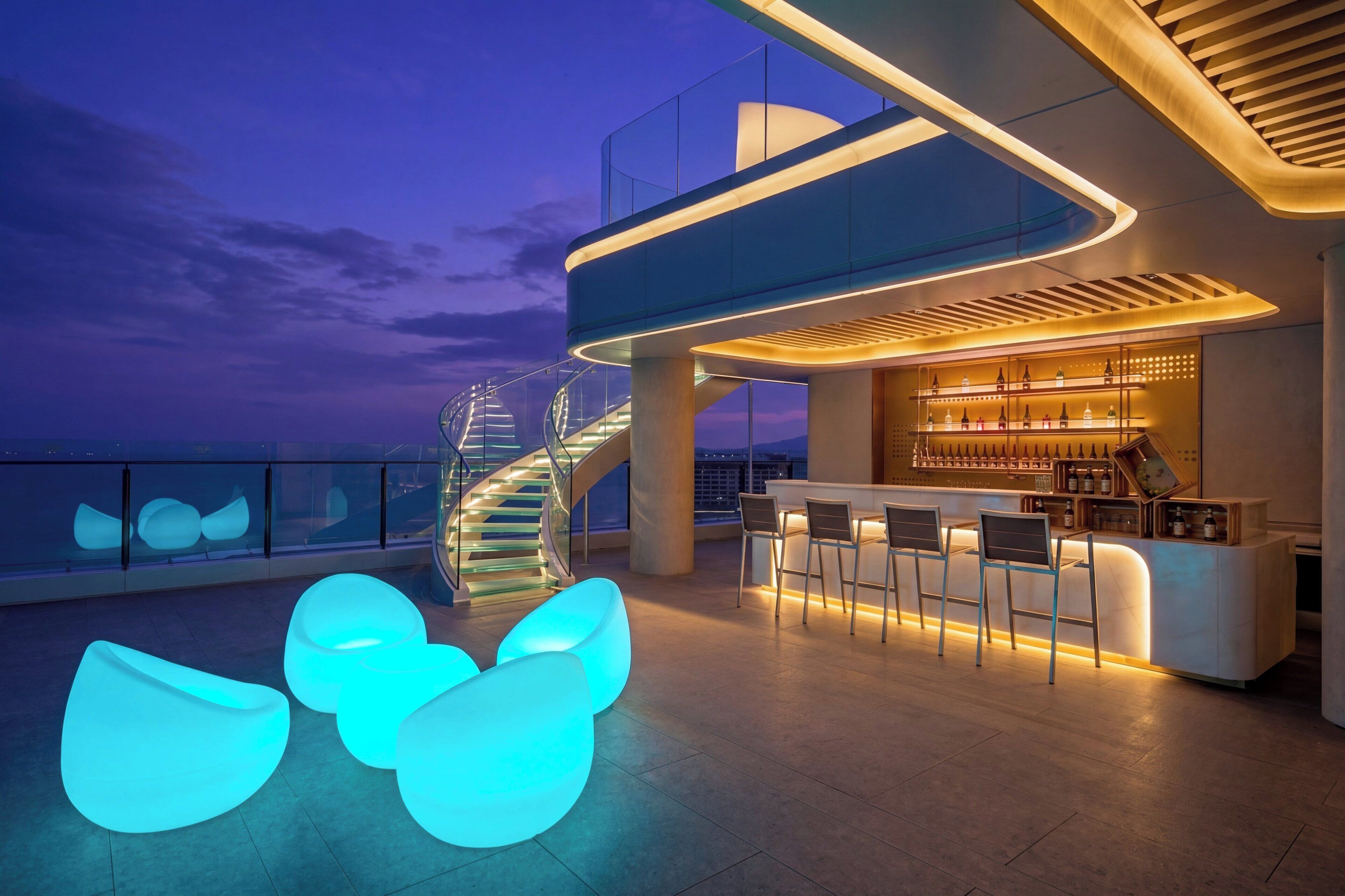 A modern bar area with a spiral staircase, bar stools, and a seating area with illuminated chairs.