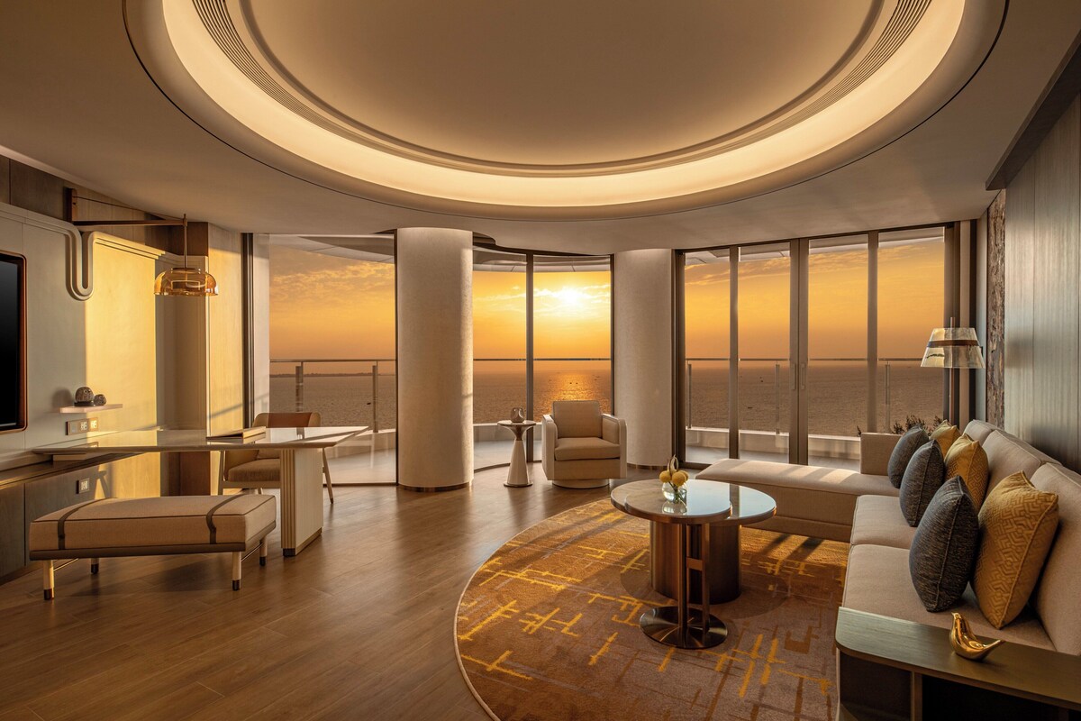 Habitación de hotel moderna con un gran ventanal que ofrece vistas al atardecer, una mesa de centro redonda, un sofá con cojines decorativos y un diseño minimalista.