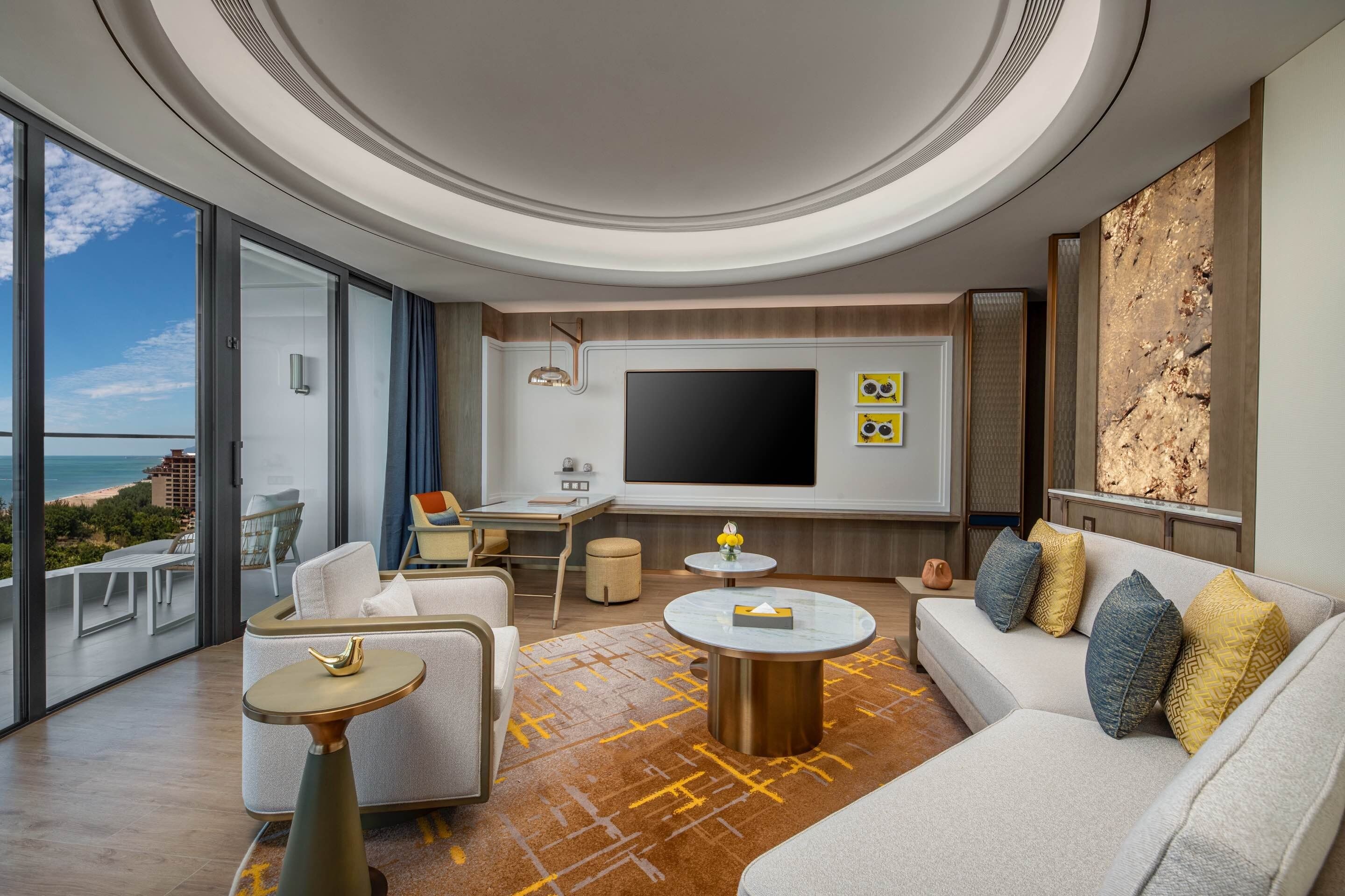 A modern living room with a large TV, comfortable seating, and a view of the ocean.