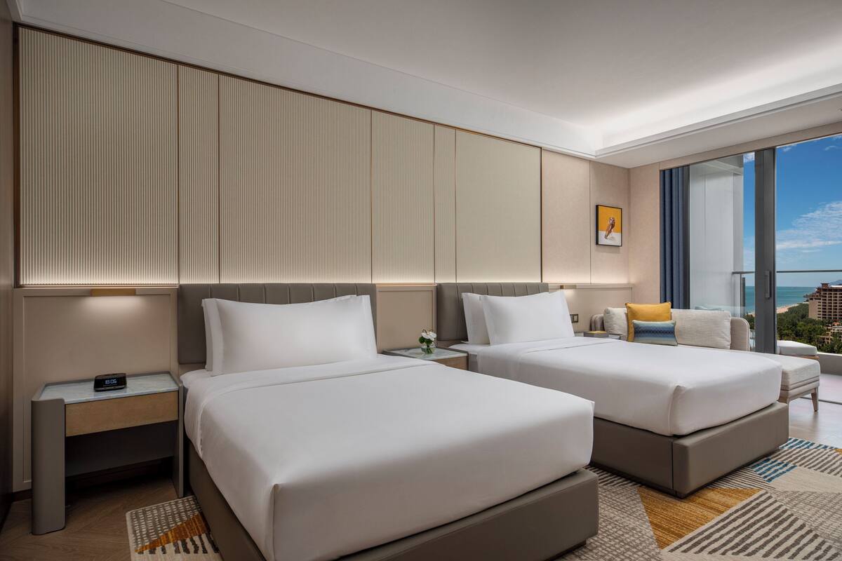 Habitación de hotel con dos camas, una mesita de noche, un escritorio, un ventanal grande con vistas a la ciudad y un diseño moderno.