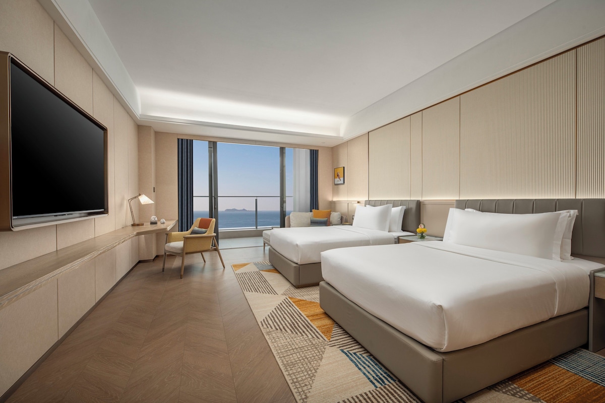 Habitación de hotel con dos camas, televisión, escritorio y vista al mar.