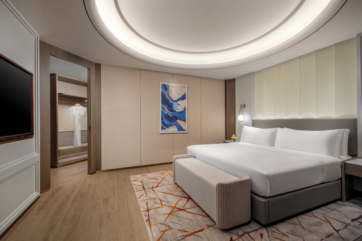 A modern hotel room with a large bed, a TV, a painting, and a bathroom with a mirror and towel rack.
