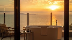 Une salle de bain moderne avec une grande baignoire, une petite table agrémentée d’objets décoratifs et une vue sur l’océan au coucher du soleil.