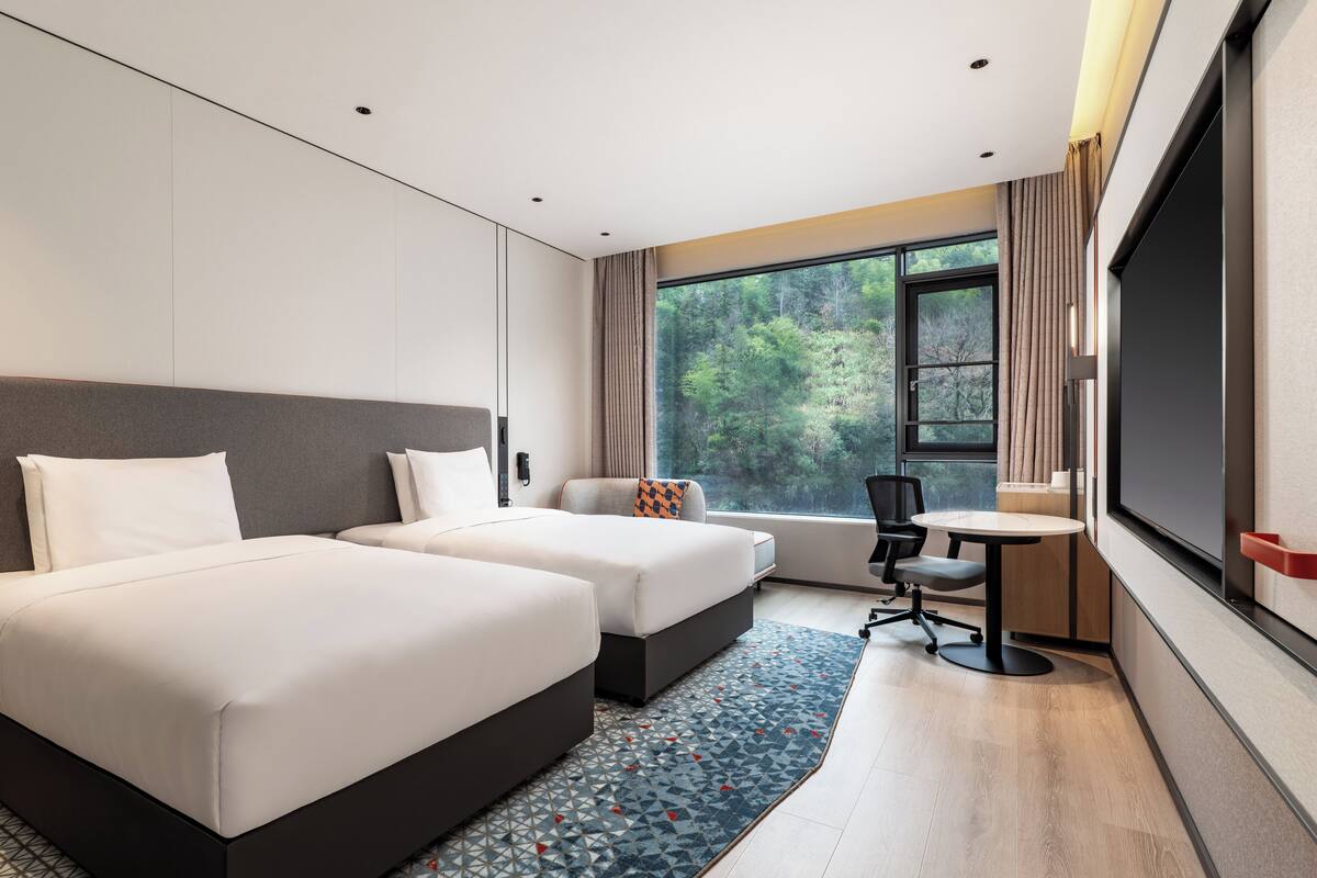 A hotel room with two beds, a desk, a chair, a TV, and a large window with a forest view.