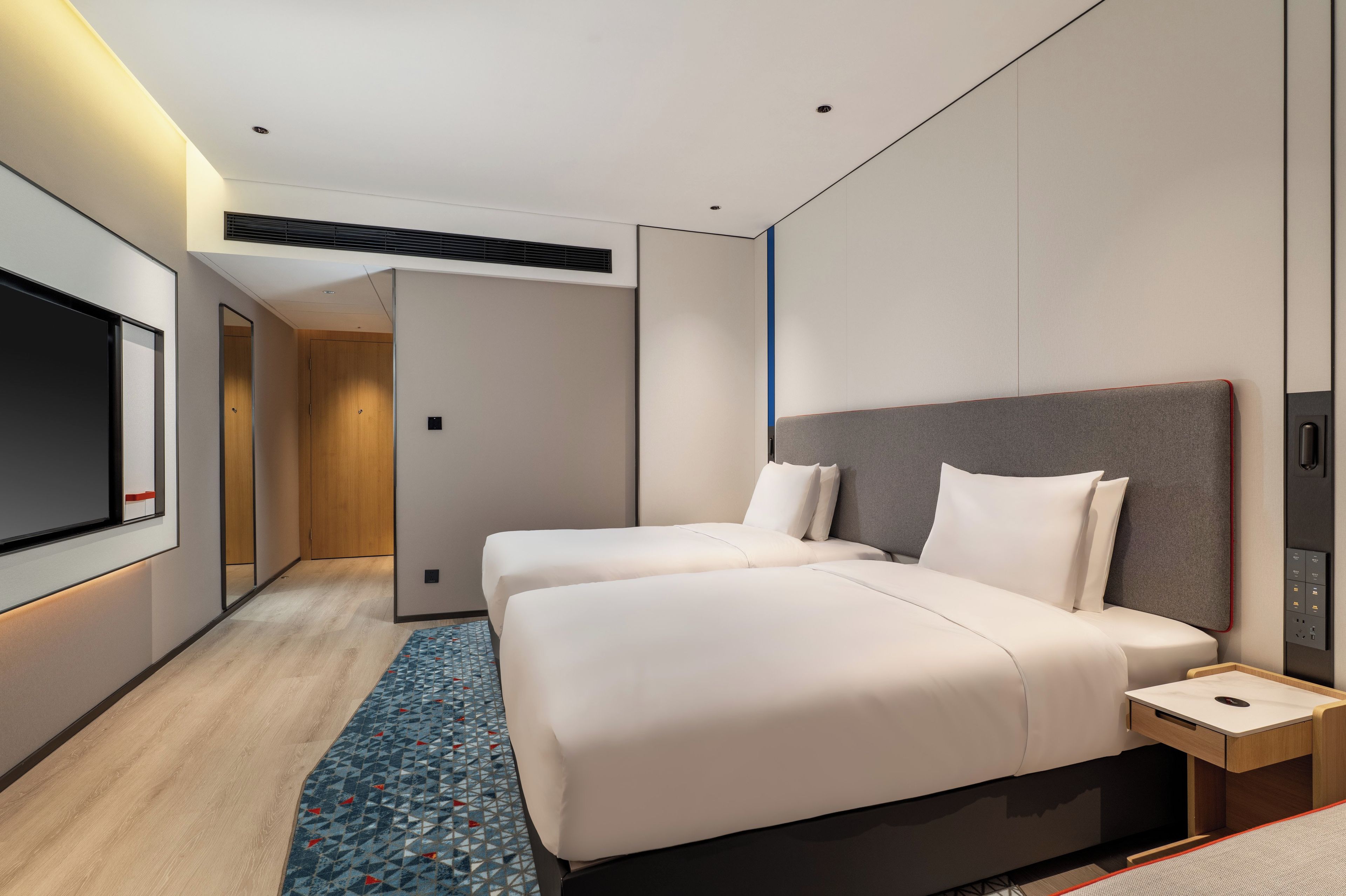 A modern hotel room with two beds, a flat-screen TV, a bedside table, and a patterned rug.