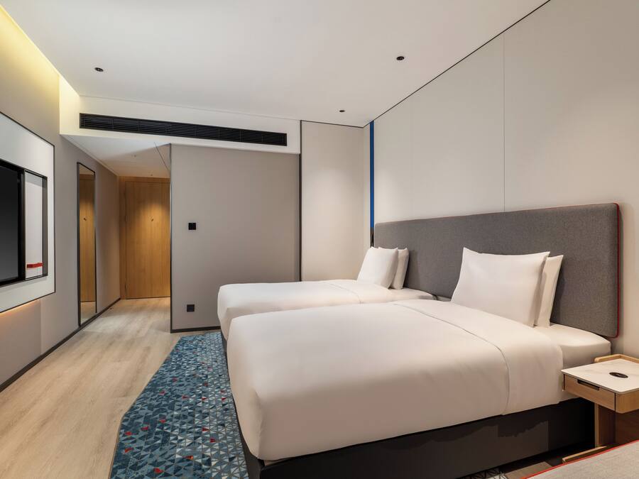 A modern hotel room with two beds, a flat-screen TV, a bedside table, and a patterned rug.