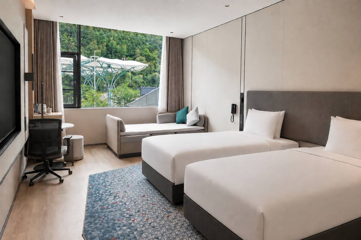 A hotel room with two beds, a desk, a chair, a TV, and a window with a view of greenery.