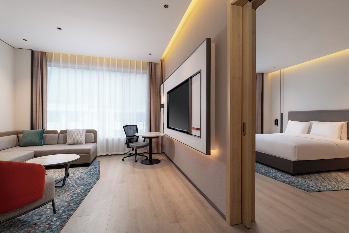 A modern hotel room with a sofa, a small table, a desk, a bed, and a TV.
