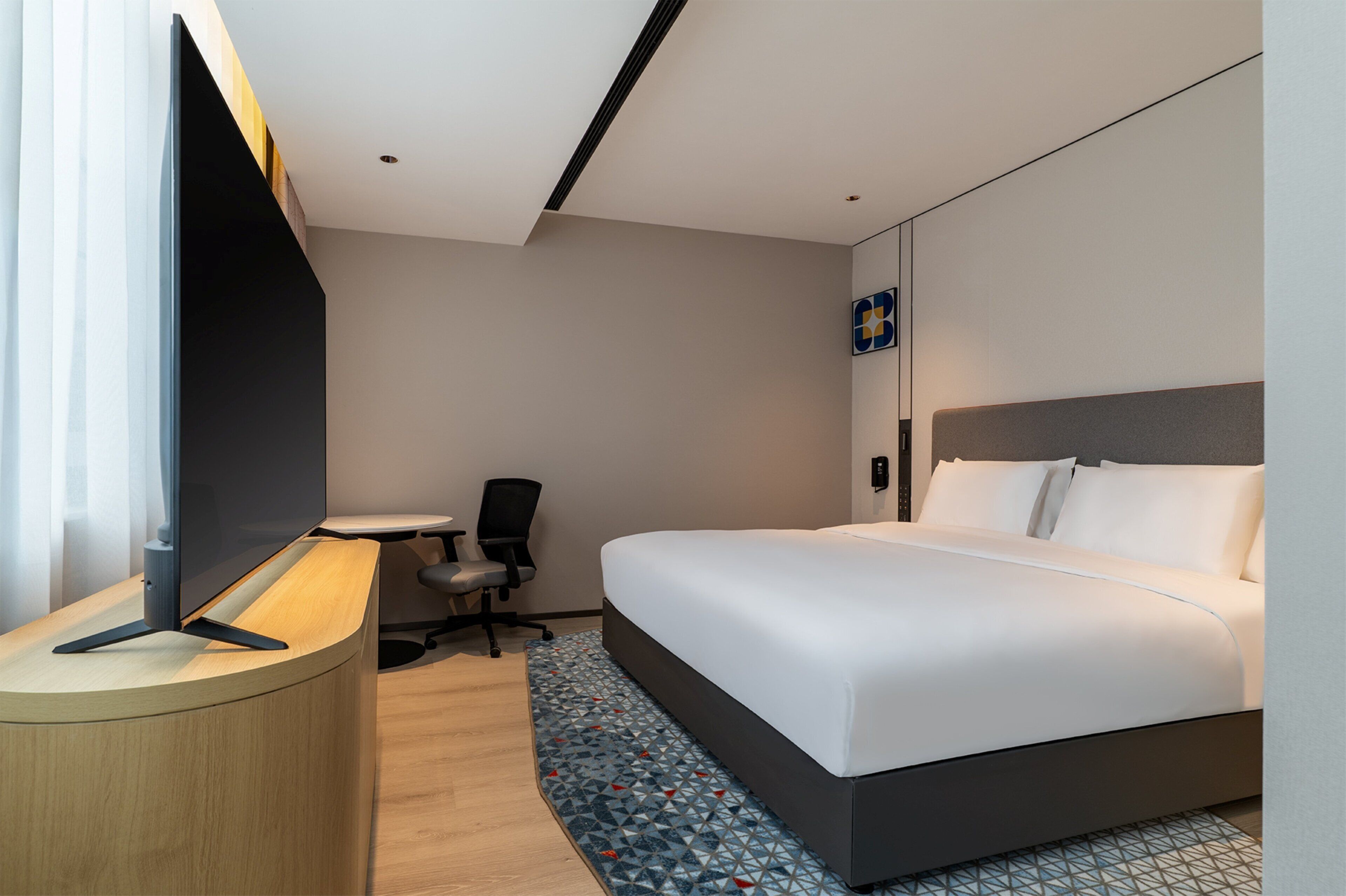 A hotel room with a bed, a desk with a flat-screen TV, a chair, and a small table.